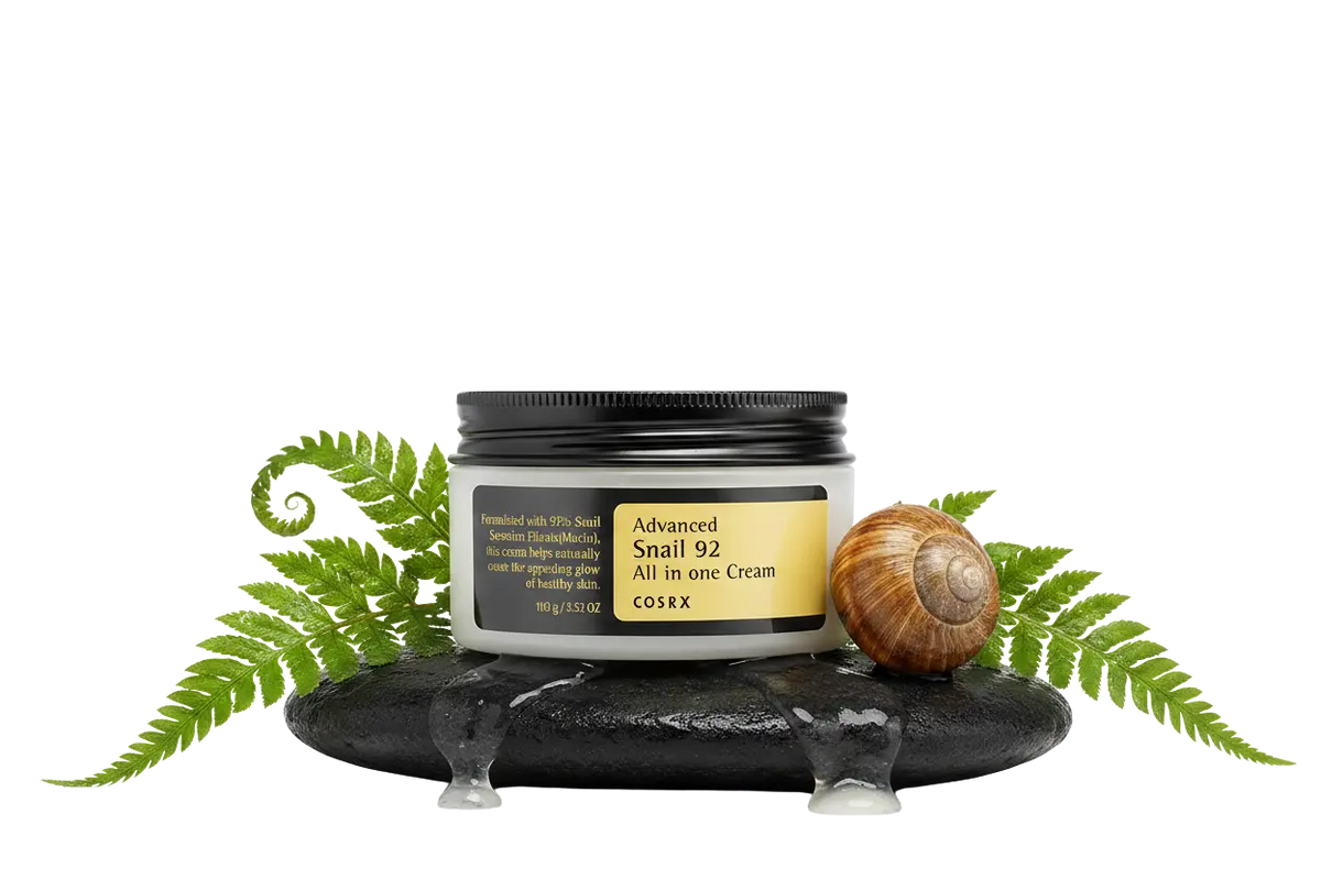 COSRX Advanced Snail 92 All in One Cream,. Hvid krukke med mørkt ribbet låg og guld label, stående på våd mørk sten omgivet af glisterende snegleslimspor, friske bregner og et sneglehus, på hvid baggrund.