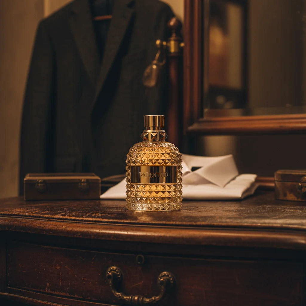 Valentino Uomo Born in Roma Gold Eau de Toilette gylden flakon på mørkt læder anretterbord med jakkesæt og hvid skjorte.